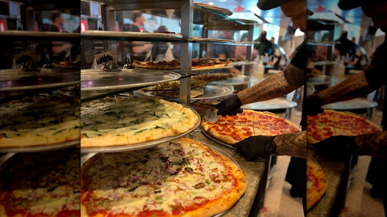 Person cutting a pizza on counter with trays and more pizzas