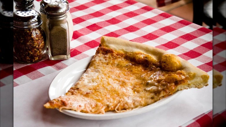 Slice of cheese pizza on a plate with checked tablecloth and pepper shakers