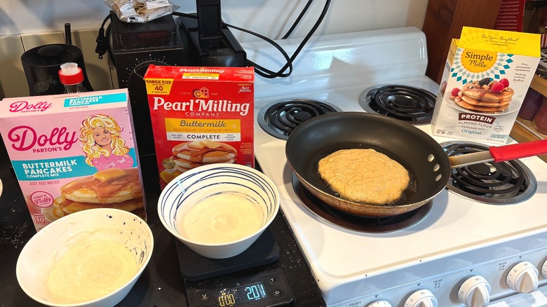 Kitchen counter and stove with pancake mix boxes, mix in bowls, and one cooking in frying pan