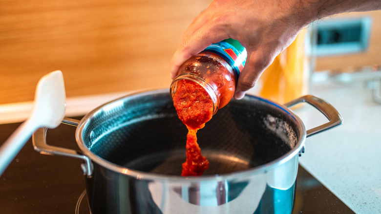 Male hand pouring tomato sauce from a jar into a large pot.