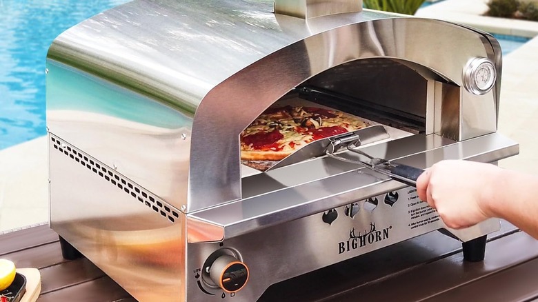 A person pulling a pizza out of a Big Horn Outdoors gas pizza oven