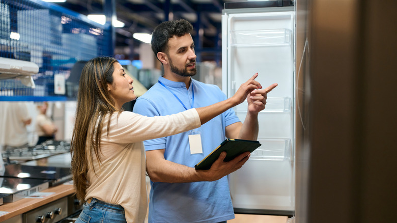 Woman pointing at refrigerator and speaking to store clerk holding electronic tablet inside appliance store