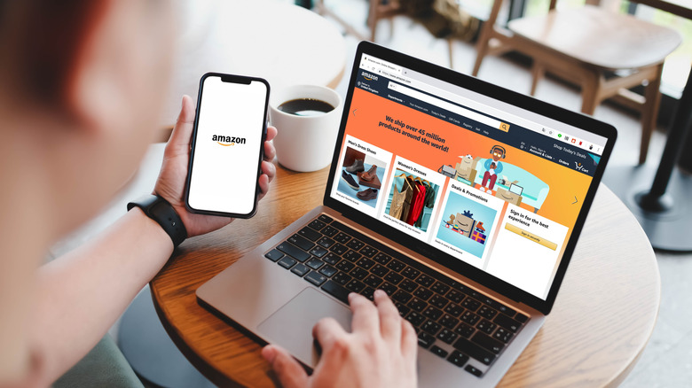 Man browsing front page of Amazon website on laptop computer and holding smartphone displaying Amazon logo