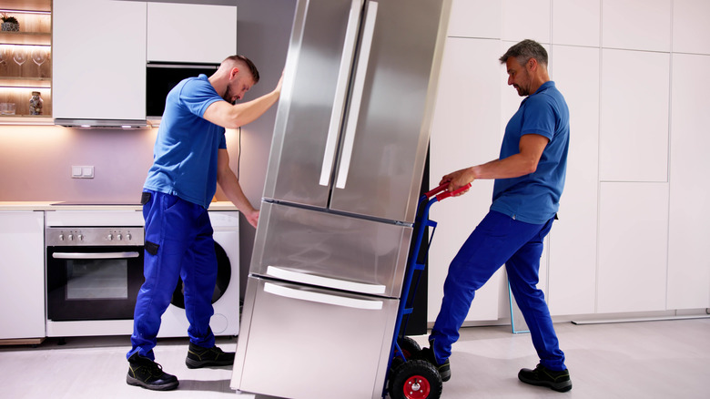 Two delivery workers moving stainless steel refrigerator into kitchen using wheeled cart