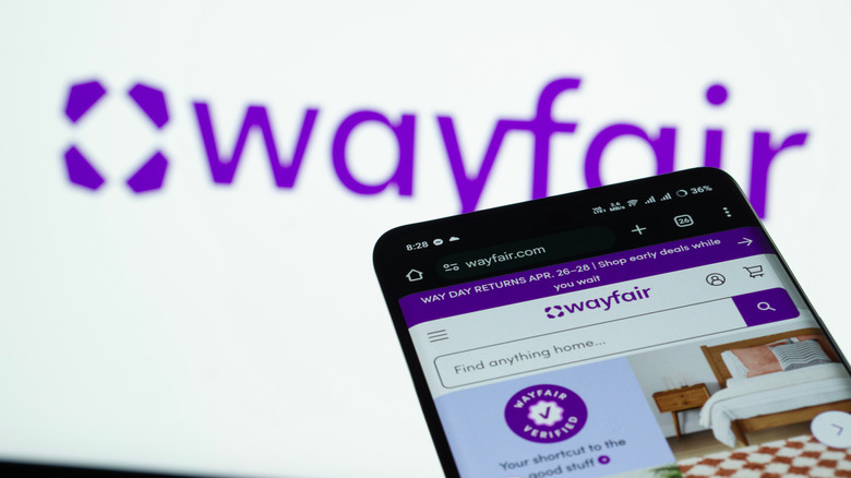 Smartphone displaying front page of Wayfair website in front of white screen displaying Wayfair logo
