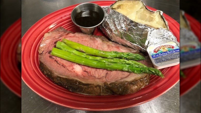 Slice of prime rib on red plate with asparagus, baked potato, and butter packet