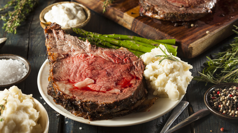 Slice of prime rib on plate with mashed potatoes, asparagus, and cutting board in background