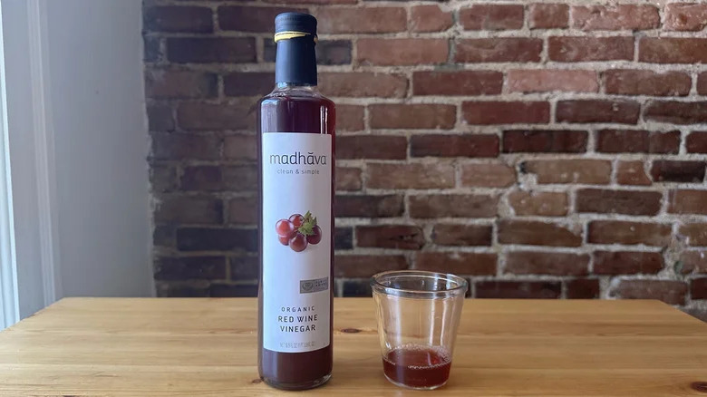 A bottle of Madhava's Organic Red Wine Vinegar sitting on a wooden table next to a glass of vinegar, with an exposed brick wall in the background