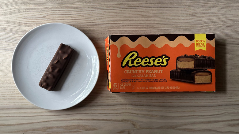 A Reese's Crunchy Peanut Ice Cream Bar sits on a white plate next to a branded package of the ice cream bars on a wooden surface