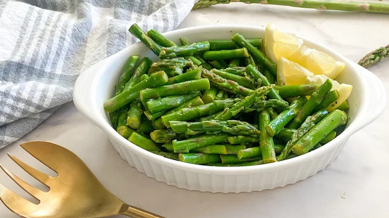 Steamed asparagus in white baking dish with lemon wedges next to a golden fork and kitchen towels