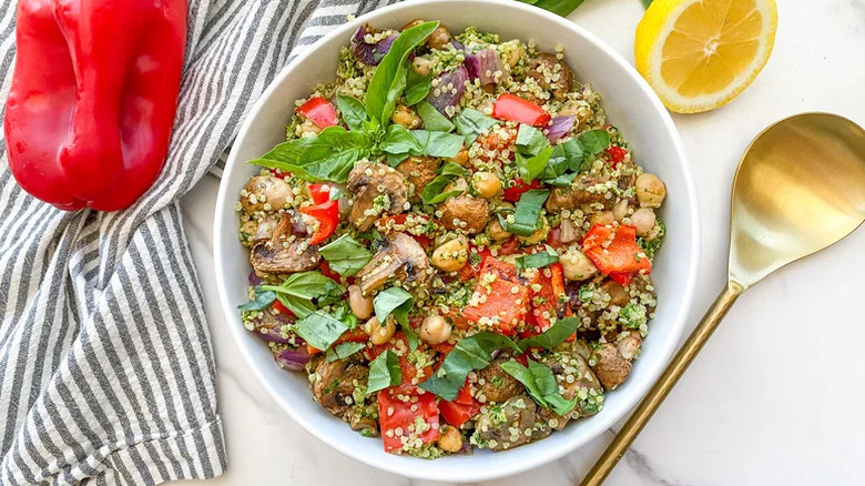 Roasted vegetable and quinoa salad in bowl, garnished with fresh basil with a golden spoon and fresh veggies and lemon on the side