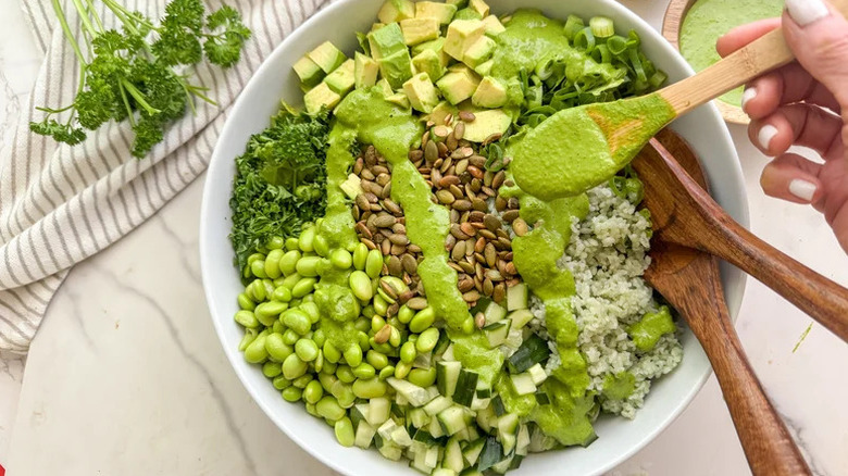 Person drizzling green dressing over a bowl of green grain salad with avocado, edamame, and cucumbers