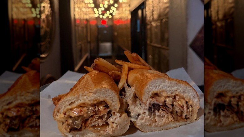 Halved chicken sandwich slathered with cheese and aioli, on a white plate with fries at the beginning of a long hallway