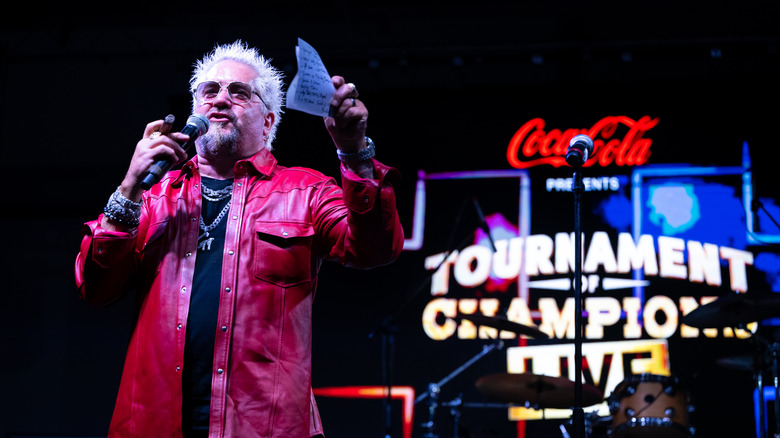 Guy Fieri onstage wearing a red leather jacket, holding a microphone and a piece of paper in front of a neon sign