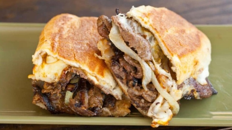 Juicy-looking steak sandwich with grilled onions and cheese, cut in half and sitting on a pea-green tray