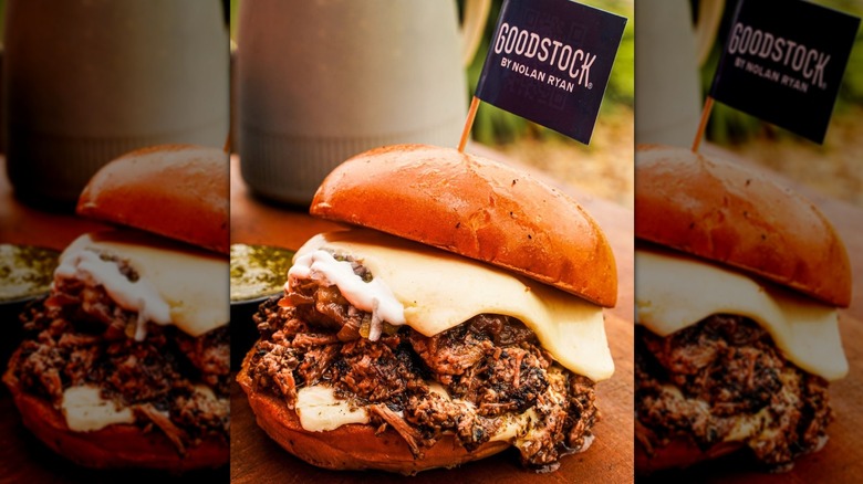 Brisket sandwich with melting cheese on a bun with a miniature blag toothpick flag that reads "Goodstock by Nolan Ryan" sticking out of it