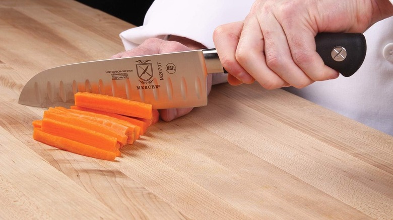 A chef cutting carrots with a Mercer Culinary santoku knife on a wooden cutting board