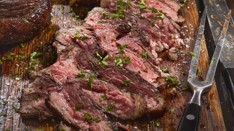 Sliced skirt steak with herbs and utensils on a wooden cutting board.