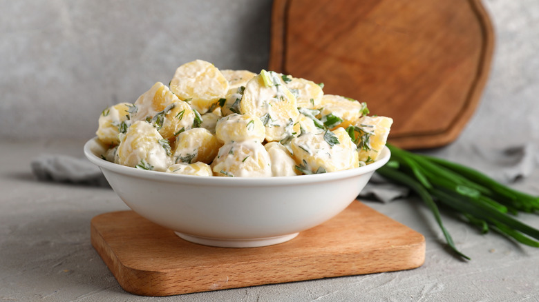 A bowl of potato salad on a wooden cutting block with greens in the background