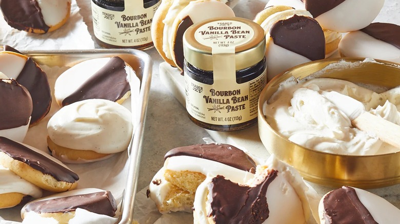 A jar of Trader Joe's Bourbon Vanilla Bean Paste sits on a counter next to trays of black and white cookies and a bowl of vanilla frosting.