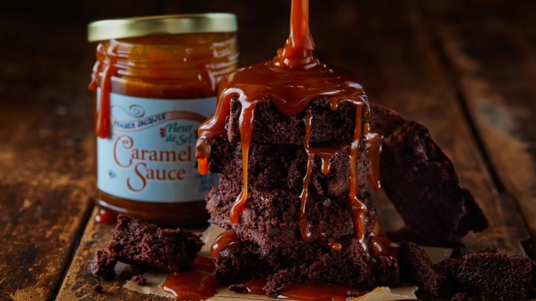 A jar of Trader Joe's Fleur de Sel Caramel Sauce sits on a wooden table behind a stack of brownies being drizzled with the sauce.