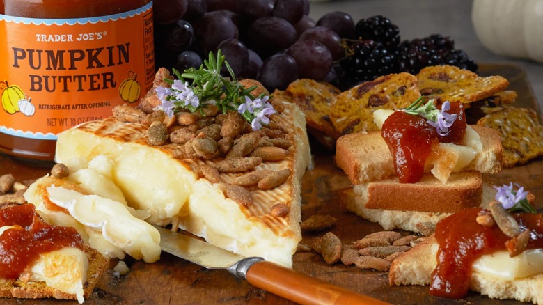 A jar of Trader Joe's Pumpkin BUtter sits on a board with soft cheese, nuts, crackers, and fruit.