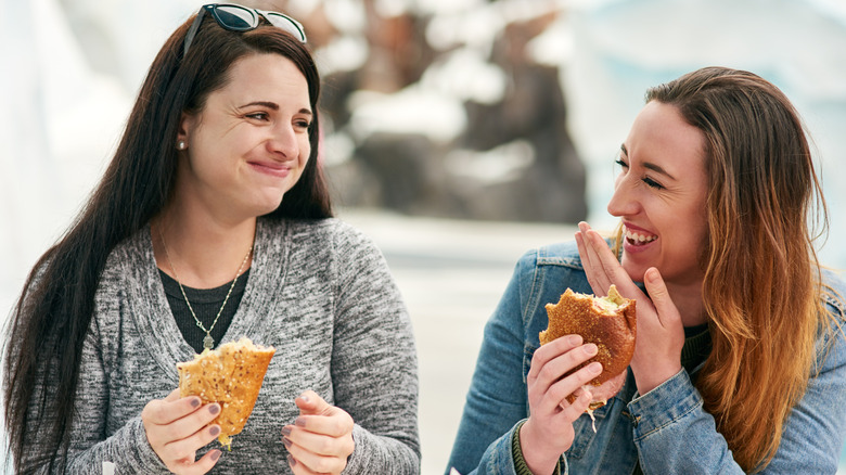 Two women laughing and eating sandwiches outside