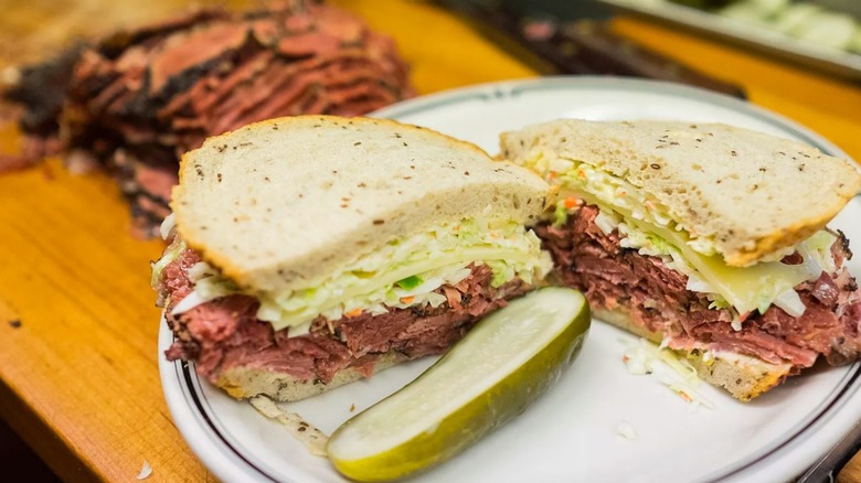 Langer's pastrami sandwich on a plate with pickle and pile of sliced pastrami background