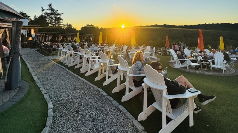 Sun dipping below green hills as people sit in rows of white Adirondack chairs on the lawn at Aquila's Nest Vineyards.