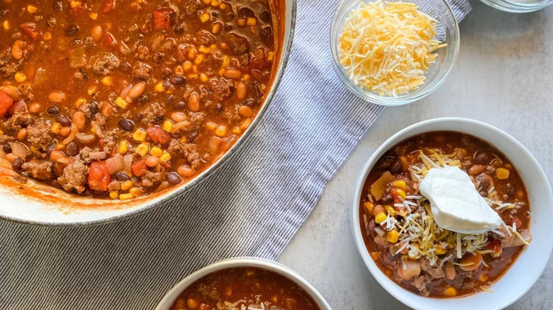 A large pot of chili sits besides two bowls of chili and a bowl of shredded cheese on a kitchen towel and an off-white surface