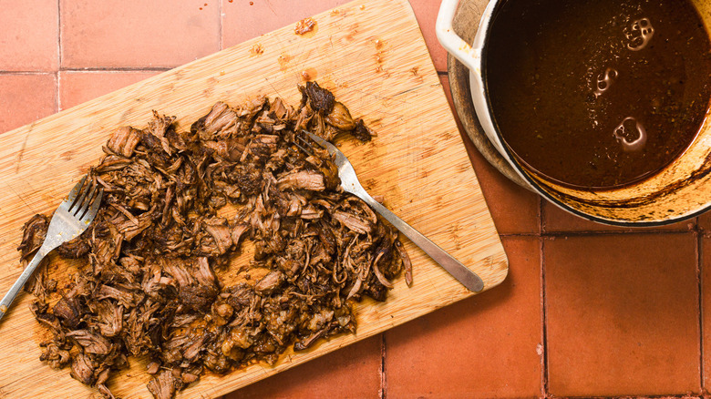 Shredding beef on cutting board besides sauce