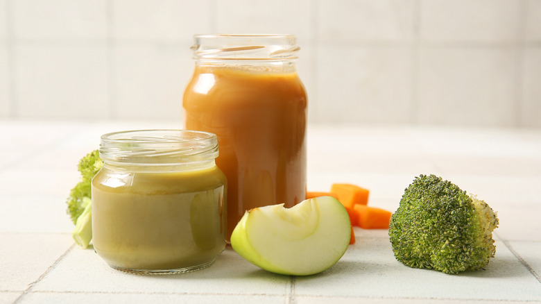 Several jars of baby food on a counter with broccoli and apples