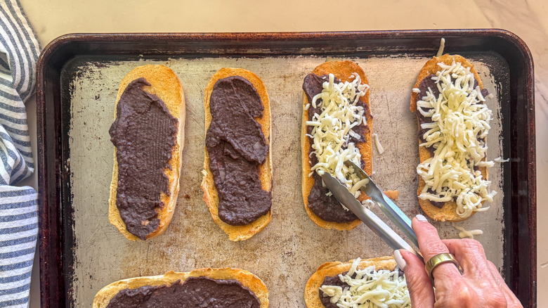 Tongs placing shredded cheese onto bread pieces with black bean puree