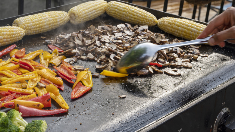 A flat-top grill with mushrooms, peppers, broccoli, and corn