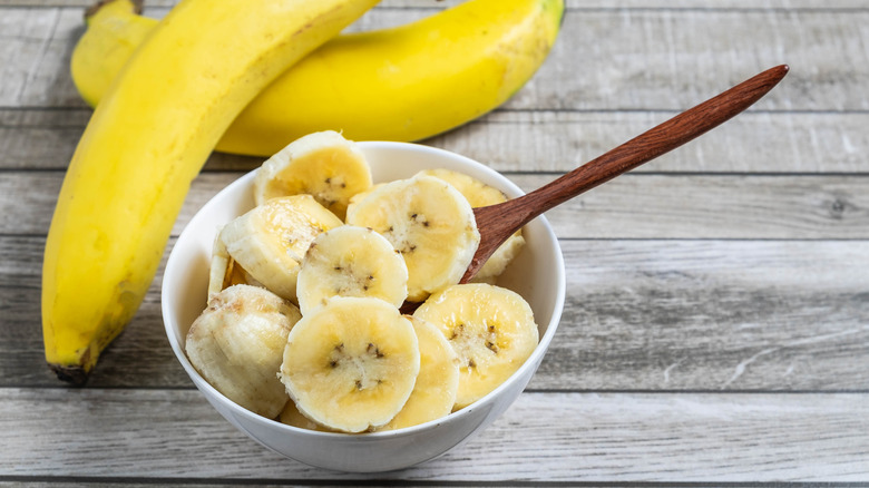White bowl of sliced bananas next to two whole bananas