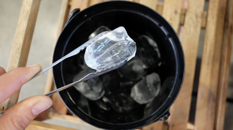 A hand holds an ice cube with tongs over bucket outside.