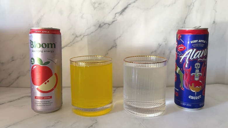 Cans of Bloom and Alani Nu apple energy drinks next to drinks in glasses on marble surface