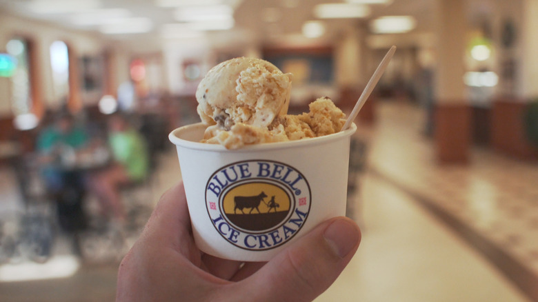 Hand holds paper cup of Blue Bell ice cream, inside shop