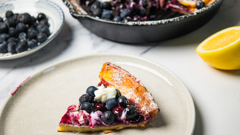 A slice of a blueberry lemon dutch baby pancake on plate, with the main pancake behind in cast iron pan.