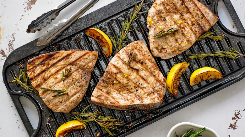 Three grilled swordfish steaks on a griddle with charred lemon and rosemary