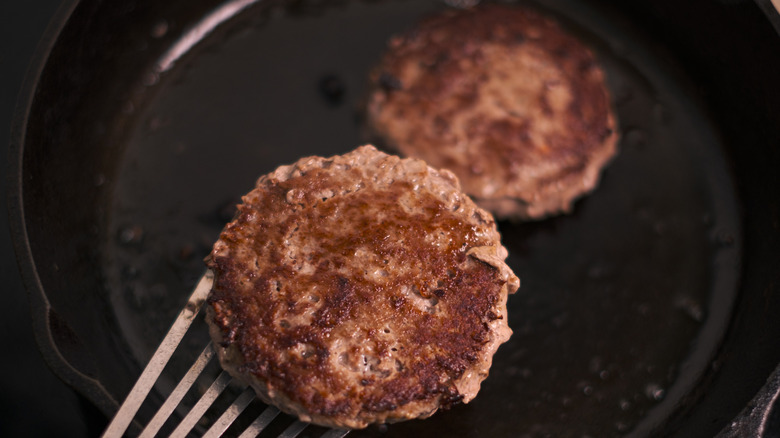 Scooping one burger patty with a spatula while the other burger cooks in a cast iron skillet