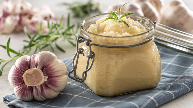 a glass jar full of garlic paste surrounded by garlic and herbs