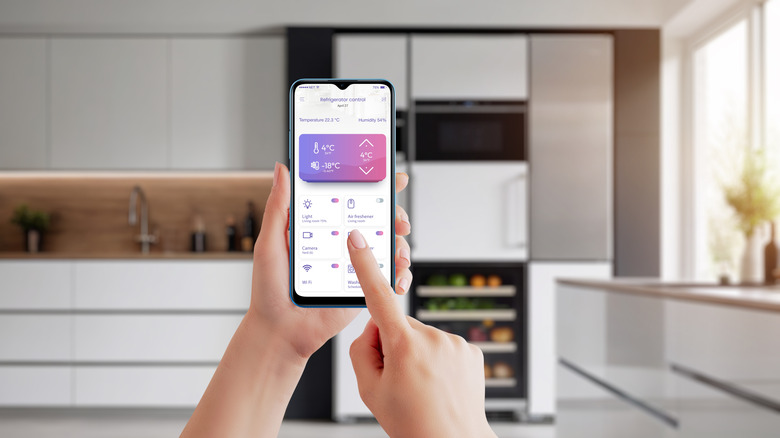 A woman's hands holding a smartphone and using an appliance-control app, with a very modern-looking kitchen visible in soft focus in the background
