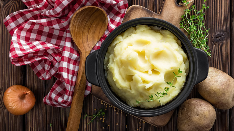 A bowl of mashed potatoes on a wooden board, with potatoes, a cloth, and spoon on the side