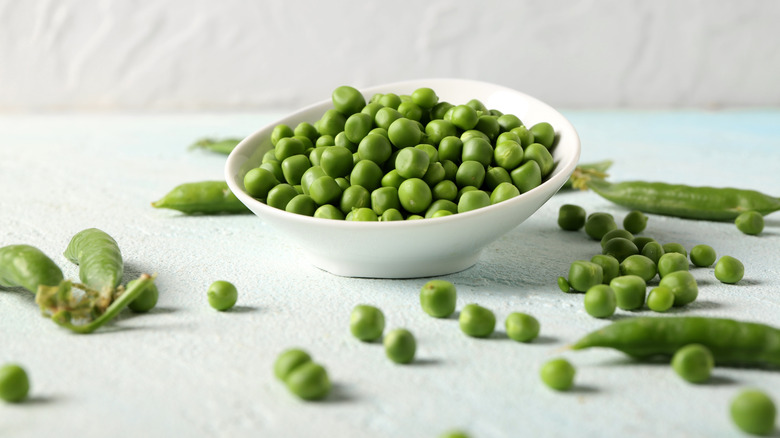 green peas in a white dish with pods strewn around a white backdrop