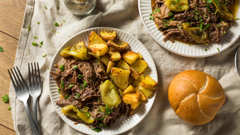Two plates of Mississippi pot roast with potatoes, next to a bread roll and two forks