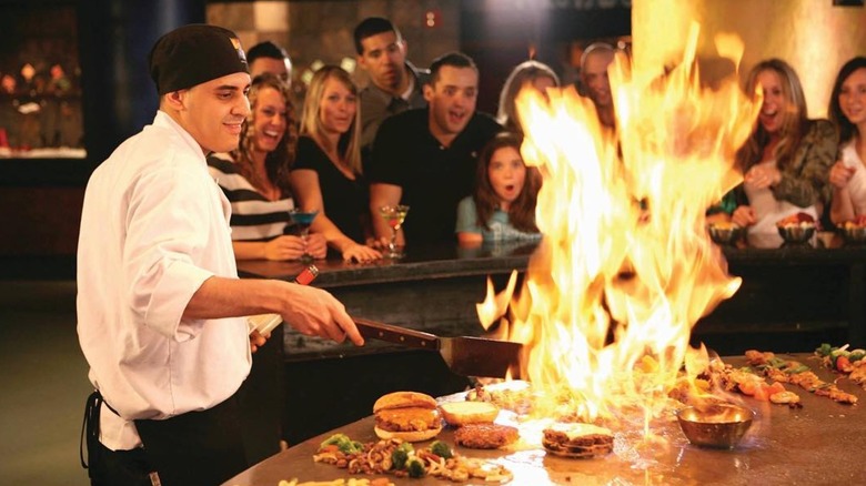 A chef prepares food on a fiery hibachi grill while customers watch