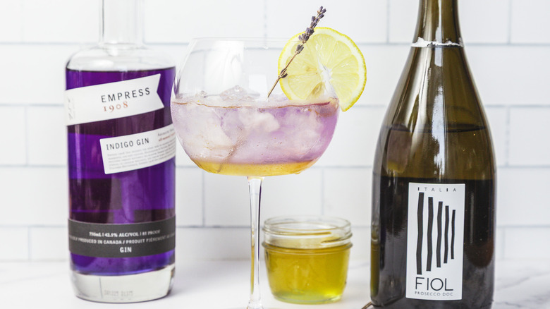 A glass of layered lavender spritz cocktail, with gin and prosecco in background.