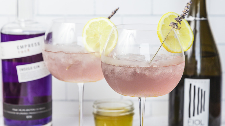 Two glasses of mixed lavender spritz cocktail, with gin and prosecco in background.