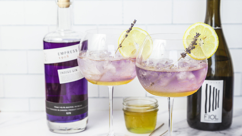 Two glasses of layered lavender spritz cocktail, with gin and prosecco in background.